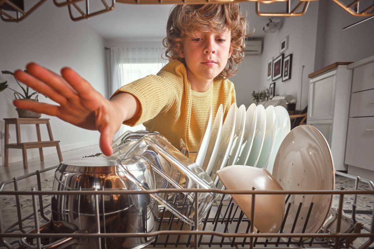 Cute boy in casual wear unloading modern dishwasher with various clean dishware during household routine in light kitchen at home