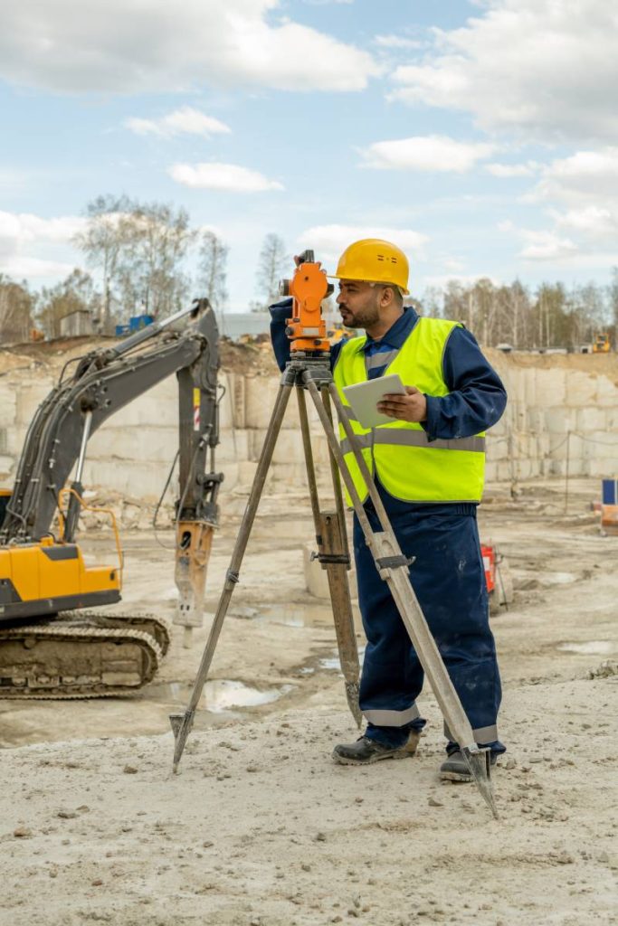 Mixed-race surveyor in workwear looking in lens of thedolite at construction site