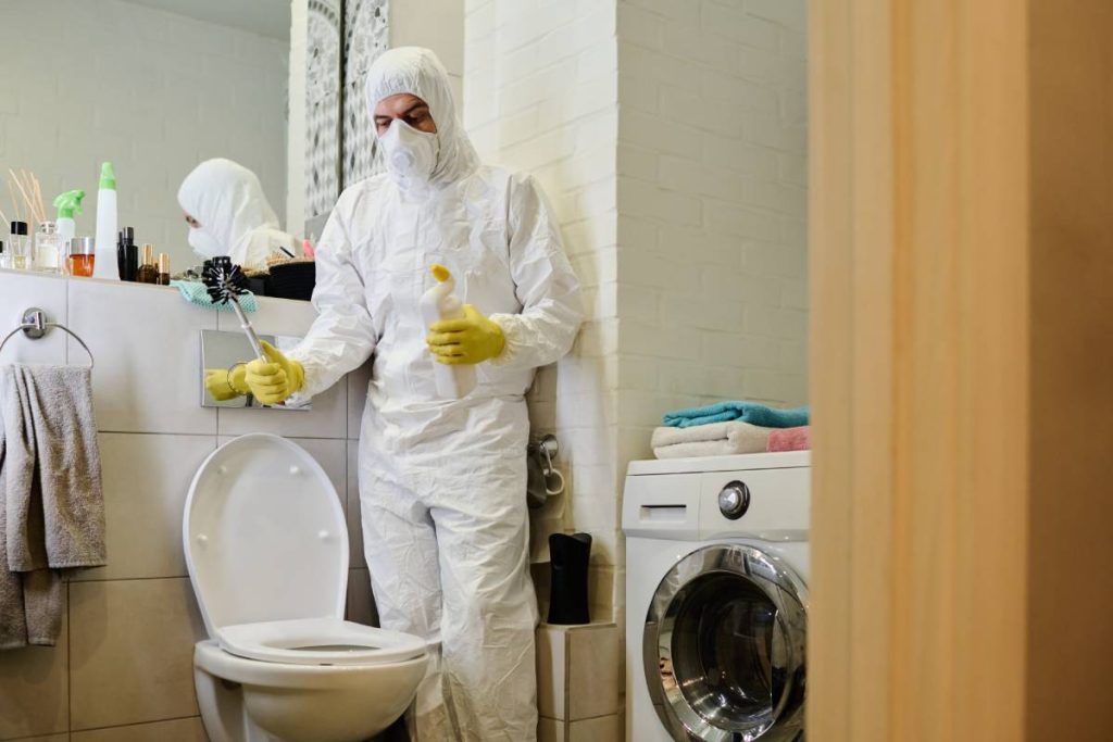 Young man in protective gloves, mask and hazmat suit cleaning toilet bowl with detergent and brush while carrying out domestic chores