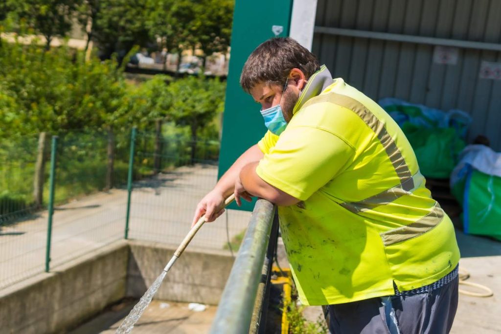 Worker in a recycling factory or clean point and garbage with a face mask and with security protections, new normal, coronavirus pandemic, covid-19. Portrait of worker cleaning with hose