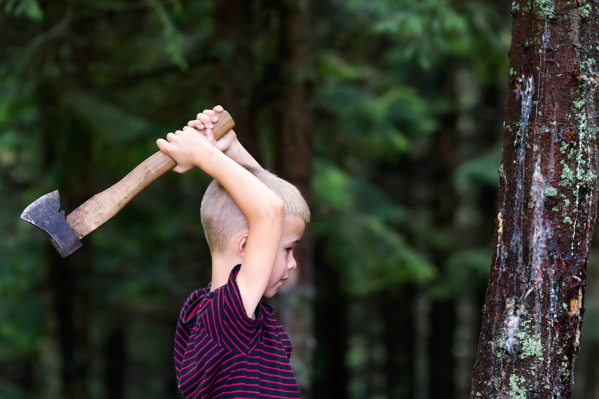 Small boy with heavy old iron axe cutting tree trump in forest on summer day. Outdoor activities and physical labor.
