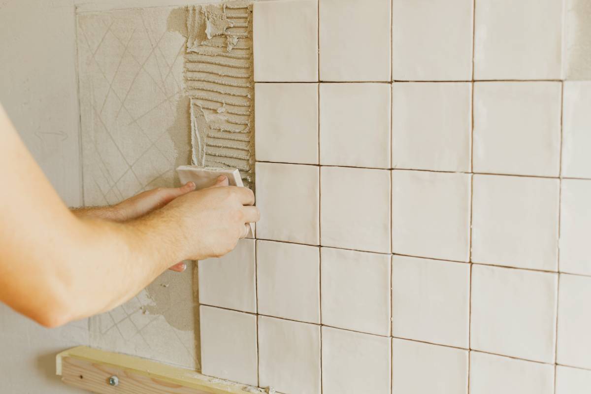 Worker installing stylish white tiles on plaster wall in sunlight. Hands laying modern square tile on adhesive close up. Kitchen or bathroom renovation, home improvement
